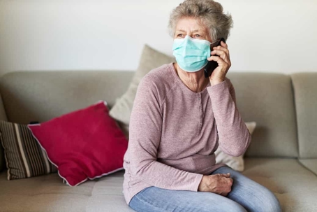 Senior woman talks on the phone while sitting on a couch wearing a face mask