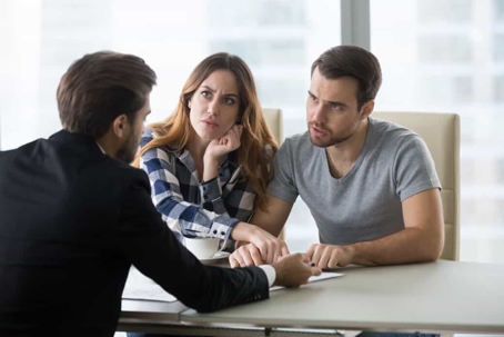 Young man wearing a suit sits at a desk in an office with a casually dressed couple as they go over paperwork