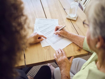 Senior woman sits at a table writing on documents with a young woman
