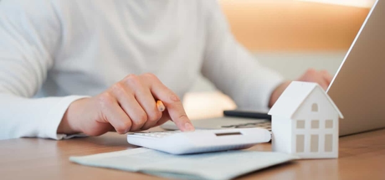 Close up of a person's hand using a calculator while working on the computer with a figurine of a house nearby