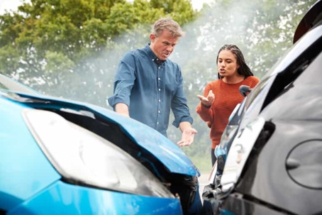 Close up of a blue car that rear-ended a black car with a man with blonde hair and a woman with braids arguing in the background