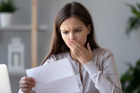 Young woman with long, brown hair covers her mouth in shock as she looks at a document she's holding
