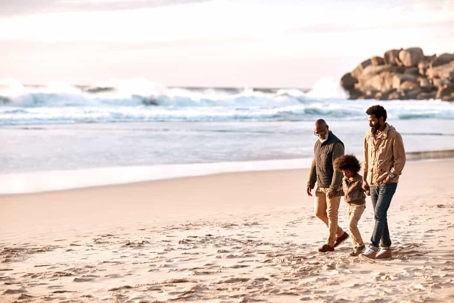 Grandfather, son, and grandson hold hands while walking on a sandy beach