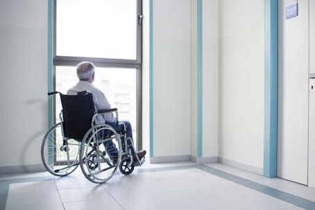 Old man with gray hair sits in a wheelchair in a medical facility staring out the window