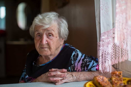 Old woman with gray hair sits with her hands folded at a table next to a window in a nursing home